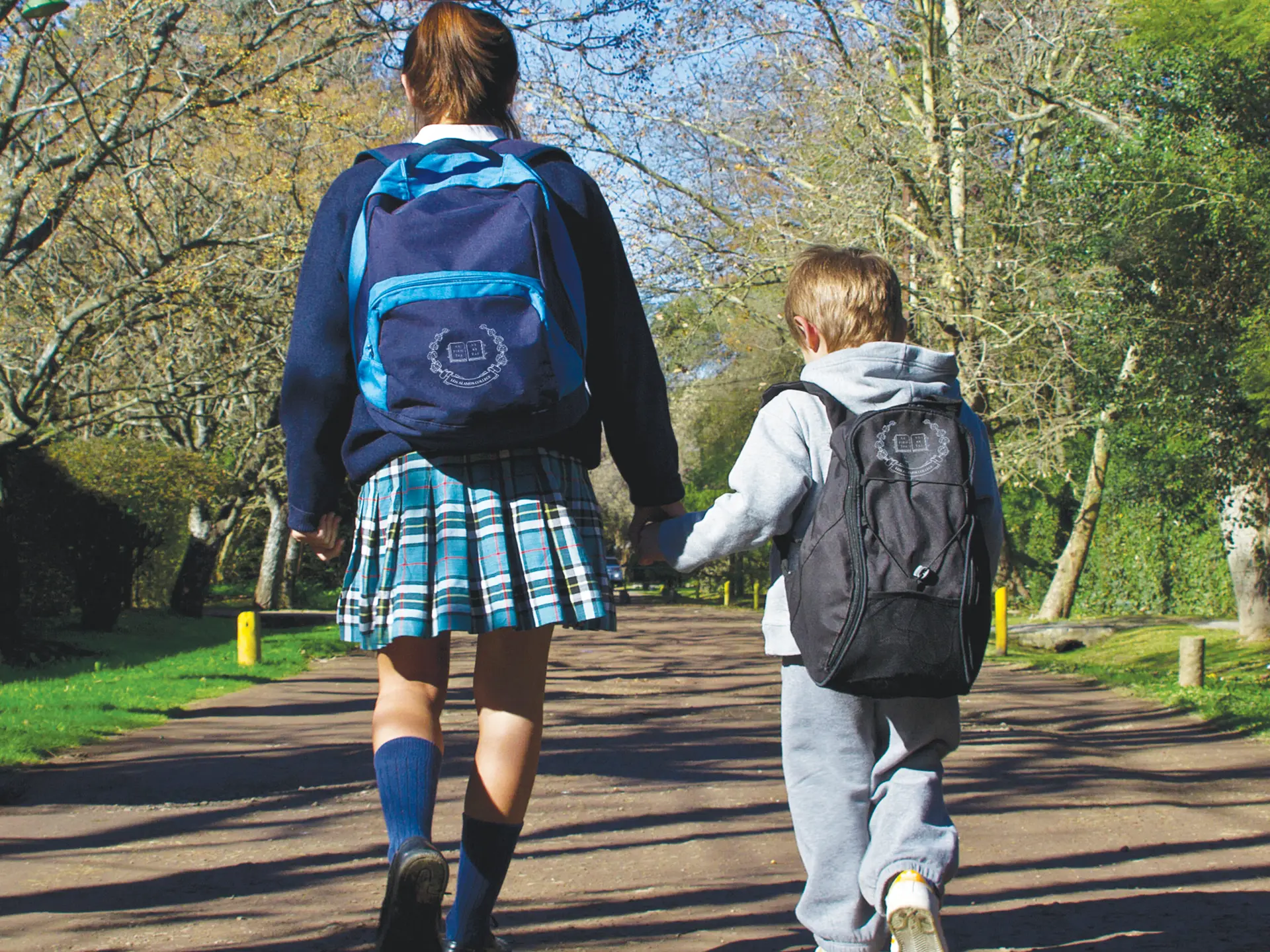 Una alumna de secundaria y un alumno pequeño caminan de la mano con mochilas del colegio por una calle arbolada
